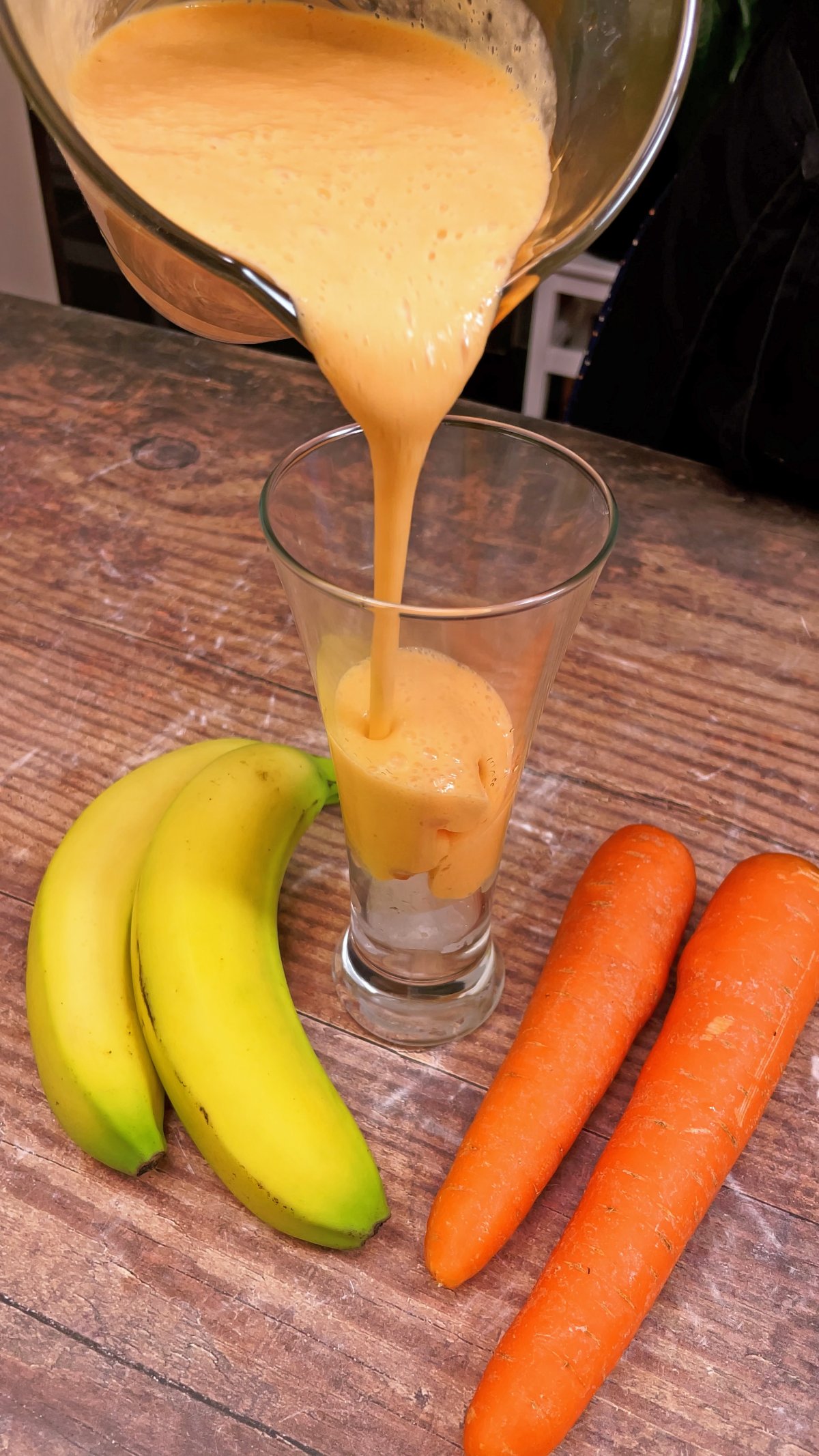 Suco de cenoura com banana sendo servido em um copo alto, com duas bananas e duas cenouras ao lado sobre uma mesa de madeira.