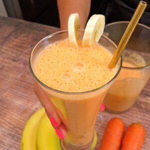 Copo de suco de cenoura com banana, decorado com rodelas de banana e um canudo dourado, sendo segurado por uma mão com unhas pintadas de rosa. Cenouras e banana