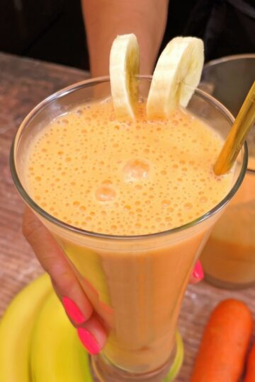 Copo de suco de cenoura com banana, decorado com rodelas de banana e um canudo dourado, sendo segurado por uma mão com unhas pintadas de rosa. Cenouras e banana